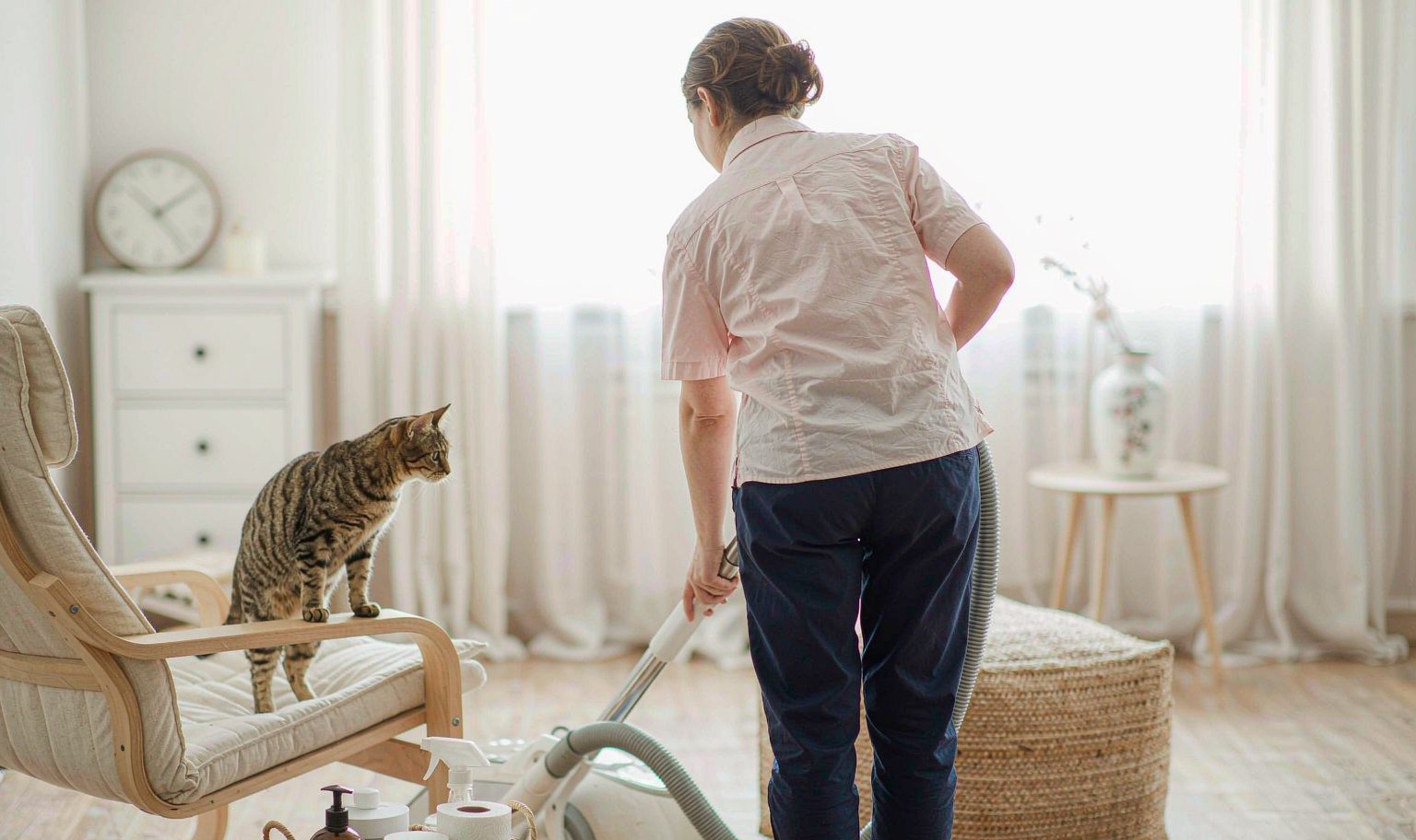 A person vacuuming a light oak floor while a cat watches from the arm of a linen armchair