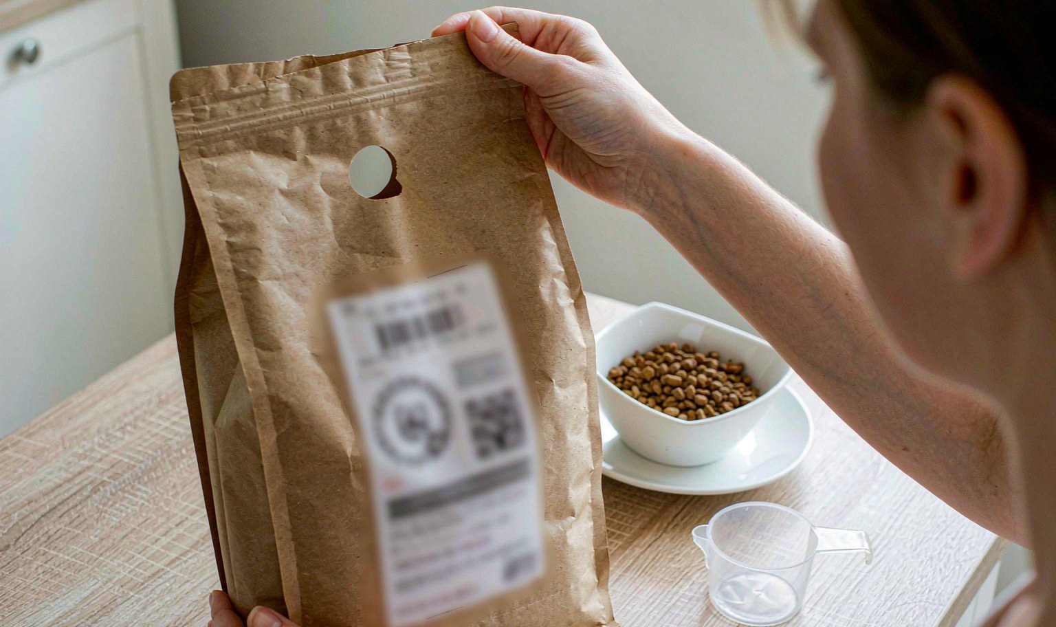 A person holding a plain bag of dry pet food to read the ingredient panel, beside a bowl of kibble and a measuring cup